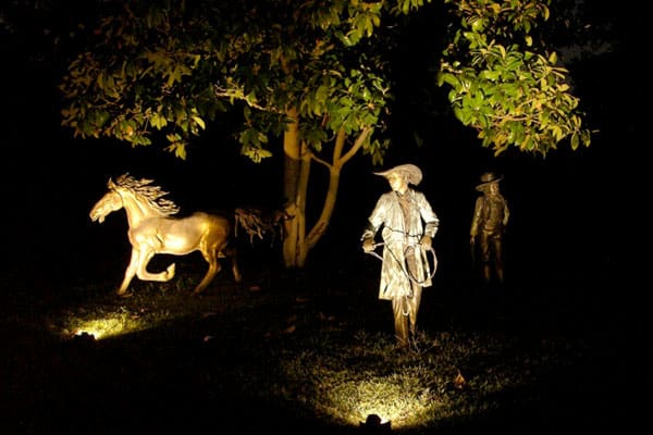 Bronze statues of a horse and cowboys are illuminated by artistic lighting