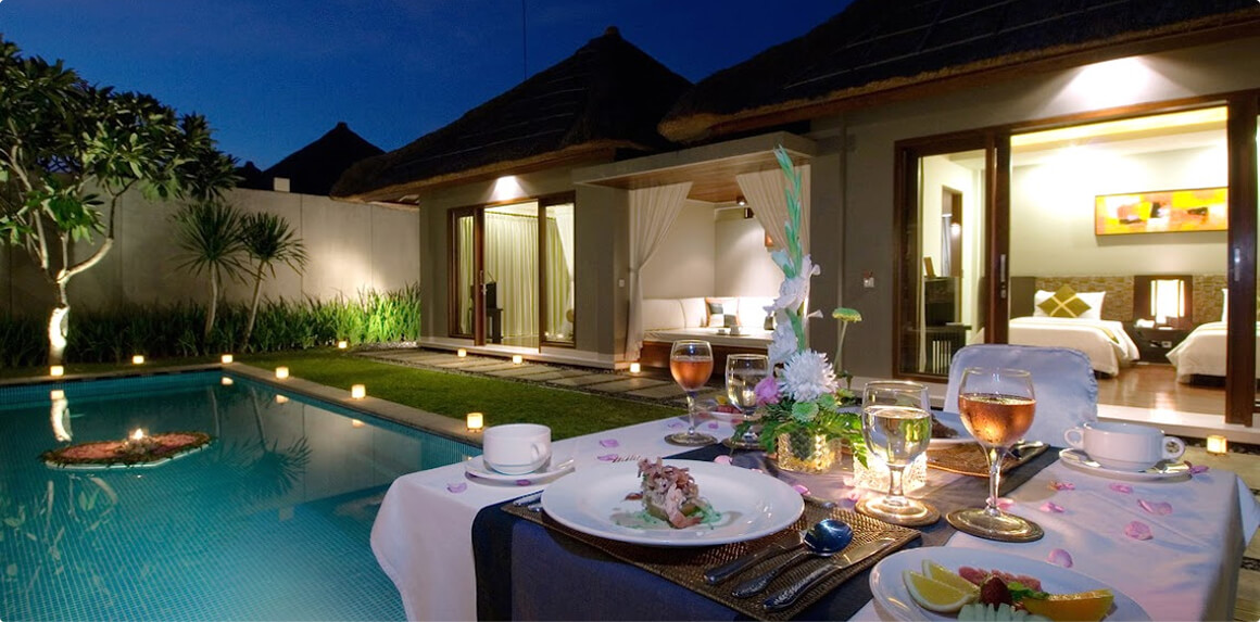 A romantic poolside dining table set for two at twilight, facing a tropical resort villa with thatched roofs and warm interior lights.