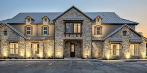 A modern two-story stone building at dusk with wooden shutters, a metal seam roof, and warm exterior lighting illuminating the front entrance and parking lot.