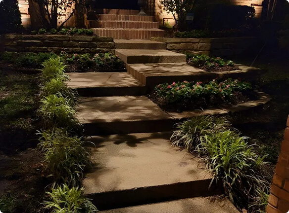 Wide concrete walkway steps illuminated at night, flanked by landscaped garden beds with flowers and ornamental grasses leading toward a brick entrance.