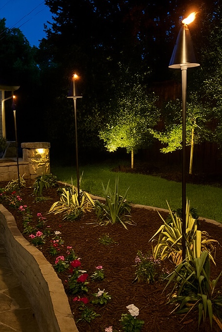 A garden bed at dusk featuring metal tiki torches with flickering flames, colorful flowers, mulch, and uplit green trees in the background.