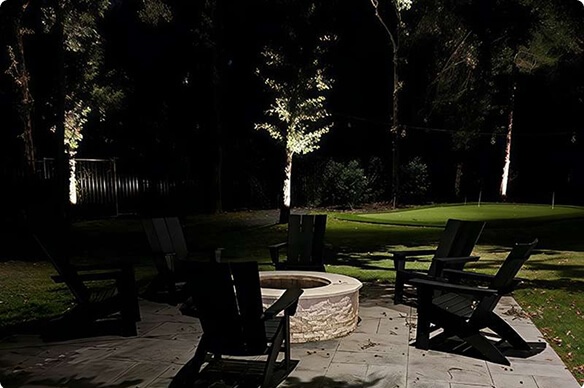 An outdoor patio at night featuring dark Adirondack chairs around a stone fire pit, with uplit trees and a glowing green lawn area in the background.