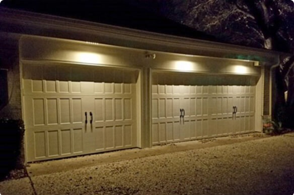 Two white, carriage-style garage doors warmly illuminated at night by downlights mounted along the soffit above a gravel driveway.