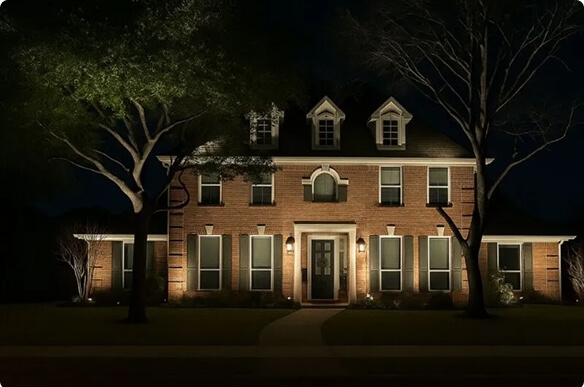 A two-story red brick home at night featuring symmetrical warm exterior uplighting, dark shutters, dormer windows, and large illuminated trees framing the front yard.