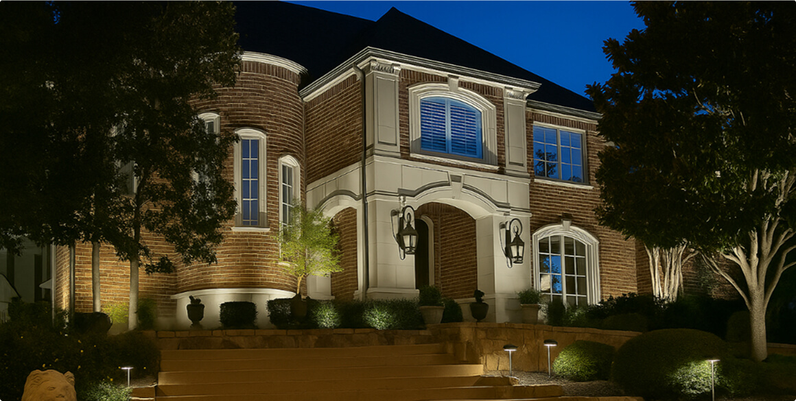 A multi-story brick estate at dusk with exterior uplighting highlighting the front steps, turret, arched stone portico, and surrounding trees.