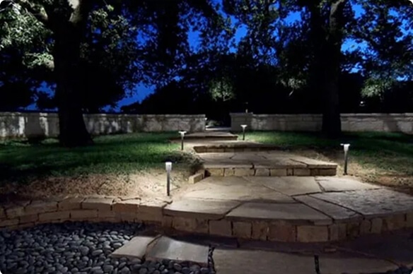 Flagstone steps leading through a grassy yard at dusk, illuminated by sleek pathway bollard lights and framed by mature trees and a stone retaining wall.