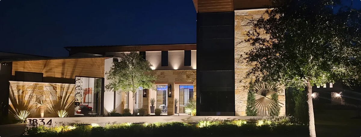 A modern architectural home illuminated at night with warm exterior uplighting on stone and wood paneling, featuring detailed landscape lighting and house numbers.