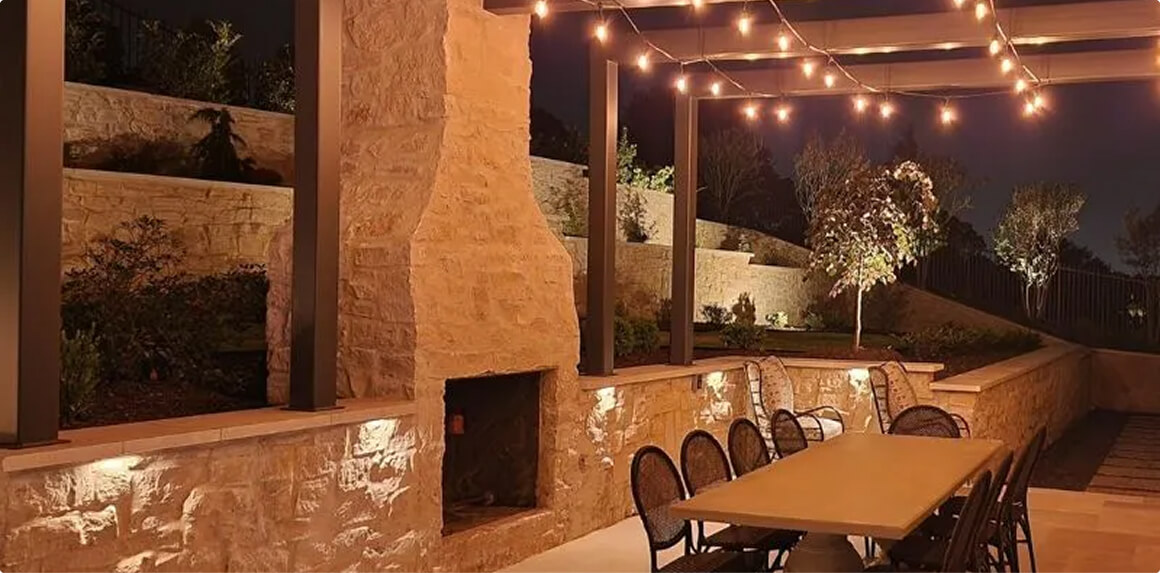 An outdoor patio dining area at night featuring a large stone fireplace, a long dining table under string lights, and a dark pergola frame.