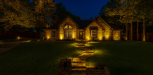 A single-story stone house at twilight, features warm landscape lighting on the facade, large surrounding trees, and a winding flagstone walkway.