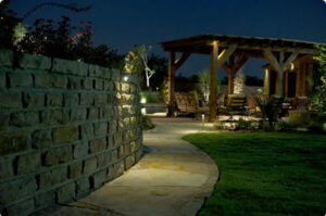 A stone walkway illuminated at night curves along a stacked stone wall toward a timber-frame pavilion with outdoor patio seating.