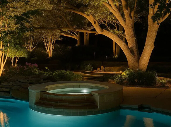 A circular hot tub integrated into a glowing blue swimming pool at night, framed by warm landscape lighting on surrounding mature trees and stone walls.