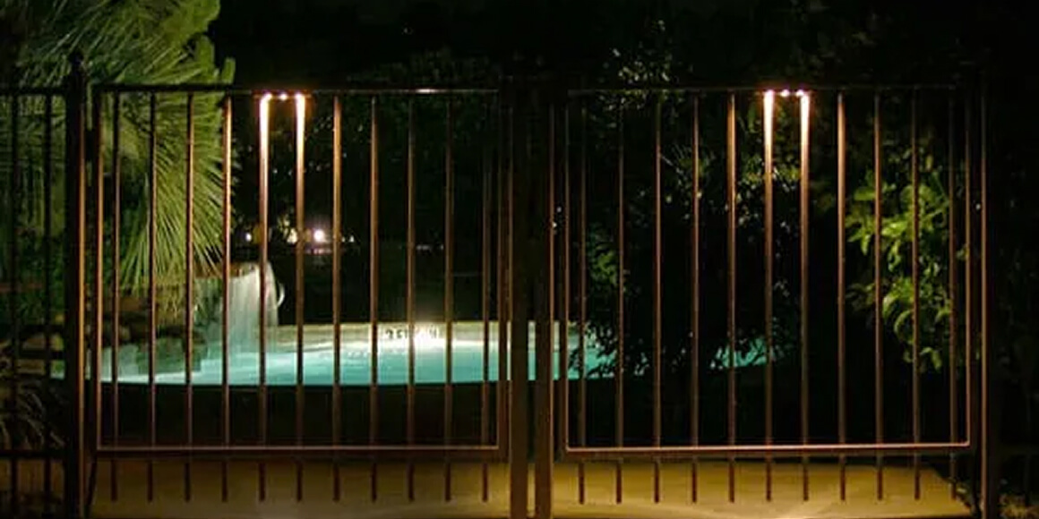 A dark wrought-iron fence gate with small integrated LED downlights overlooking an illuminated backyard swimming pool at night.