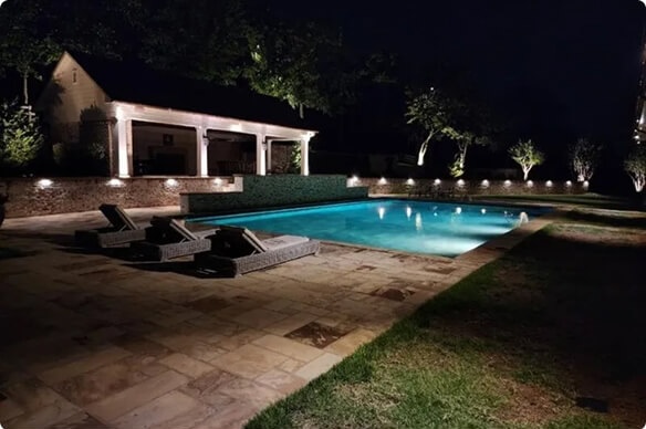 A swimming pool glowing with bright aqua water at night, featuring pool lounge chairs, an illuminated stone retaining wall, and a covered pool house in the background.