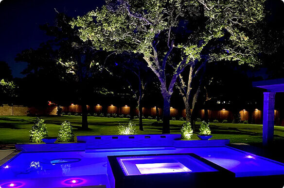 A modern backyard landscape at night featuring a swimming pool with vibrant blue and purple LED lighting, set against a lawn with uplit trees and a glowing wooden fence line.