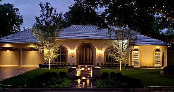A single-story stucco home at dusk featuring warm architectural lighting, uplit young trees, and illuminated stone walkway steps leading to an arched front entry.