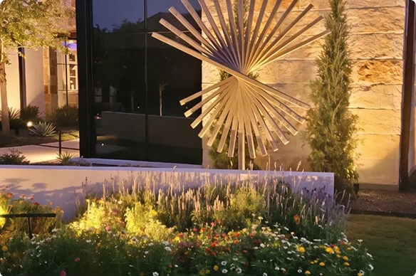A modern kinetic sunburst wind sculpture installed in a garden bed with warm landscape lighting, colorful flowers, and a stone wall backdrop at night.