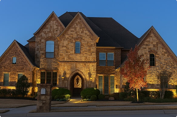 A multi-story stone home at dusk with warm exterior accent lighting highlighting the pointed arch entryway, stone facade, and autumn tree in the front yard.