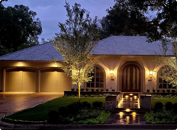 A modern single-story home illuminated at dusk with warm exterior wall sconces, uplit young trees, and glowing step lights along the front walkway.