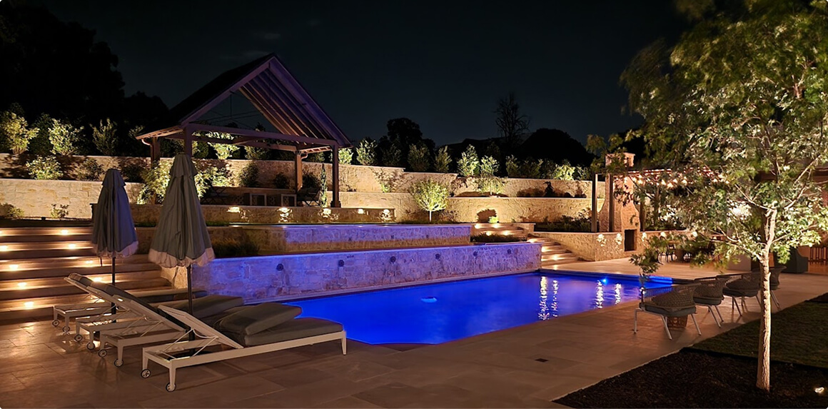 A modern backyard landscape at night featuring a swimming pool with vibrant blue and purple LED lighting, set against a lawn with uplit trees and a glowing wooden fence line.